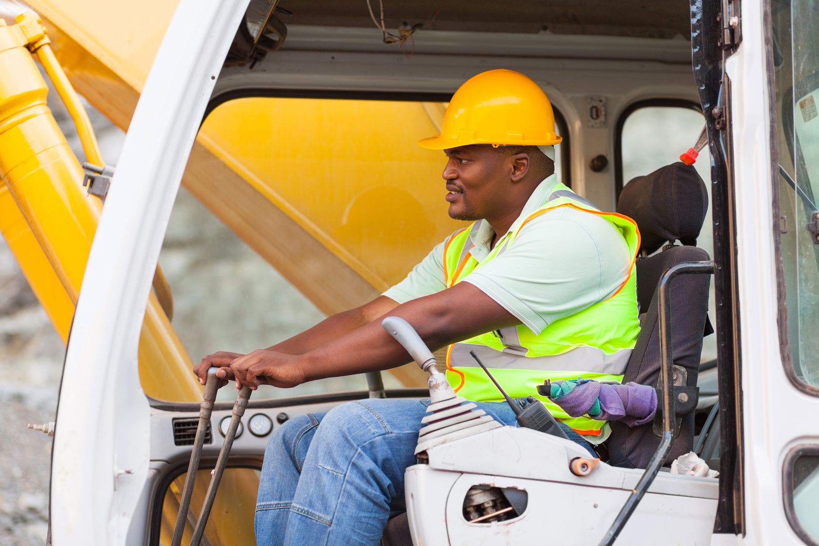 A Black male wearing a safety vest and hat operates a construction crane.