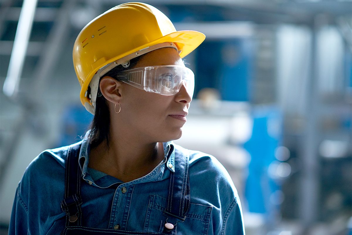 A woman wearing a yellow hard hat and safety glasses looks to the side.