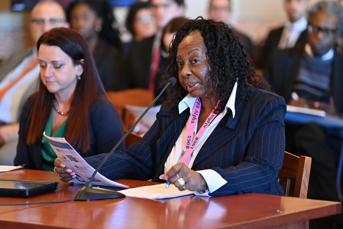 Senator Hunter is seated at a table and speaks into a microphone while holding a piece of paper and testifying before a Senate committee.